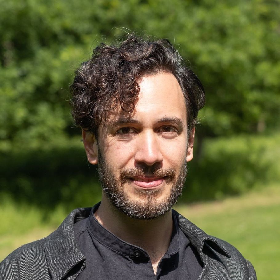 Portrait of a man outdoors in a black jacket, standing in front of green trees and sunlight