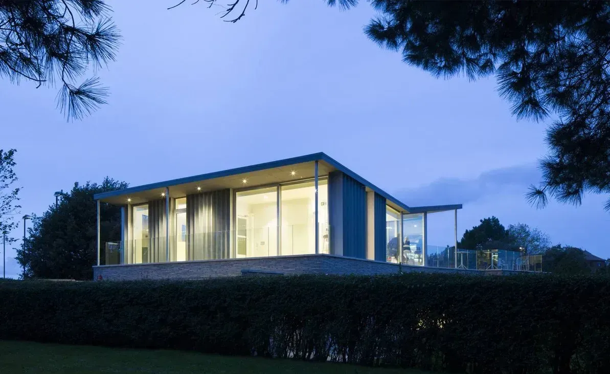 Modern glass house with warm interior lights at dusk, framed by trees and a blue evening sky