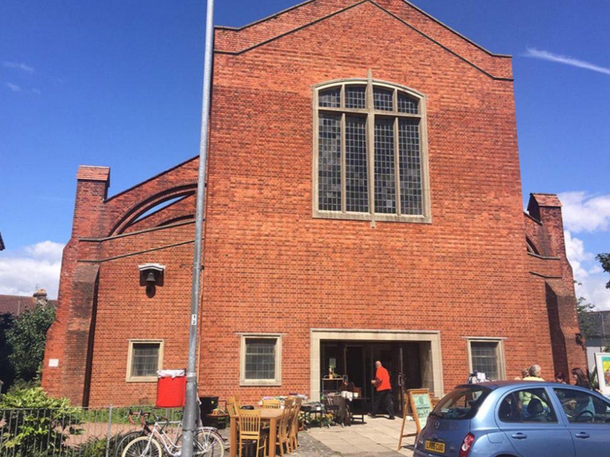 Brick building with large arched window, outdoor tables, and a parked blue car under a clear sky.