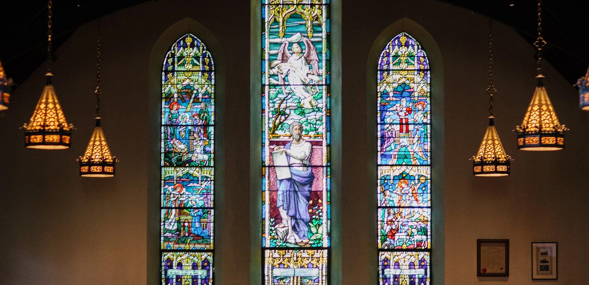 Three colorful stained-glass church windows with hanging pendant lights in a dim interior