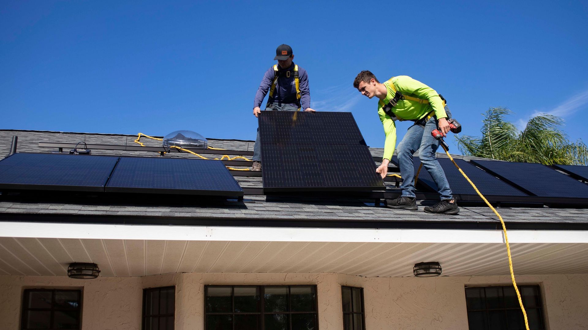Two workers installing black solar panels on a roof under a blue sky