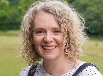 Smiling woman with curly blonde hair in a polka-dot top outdoors, with greenery in the background