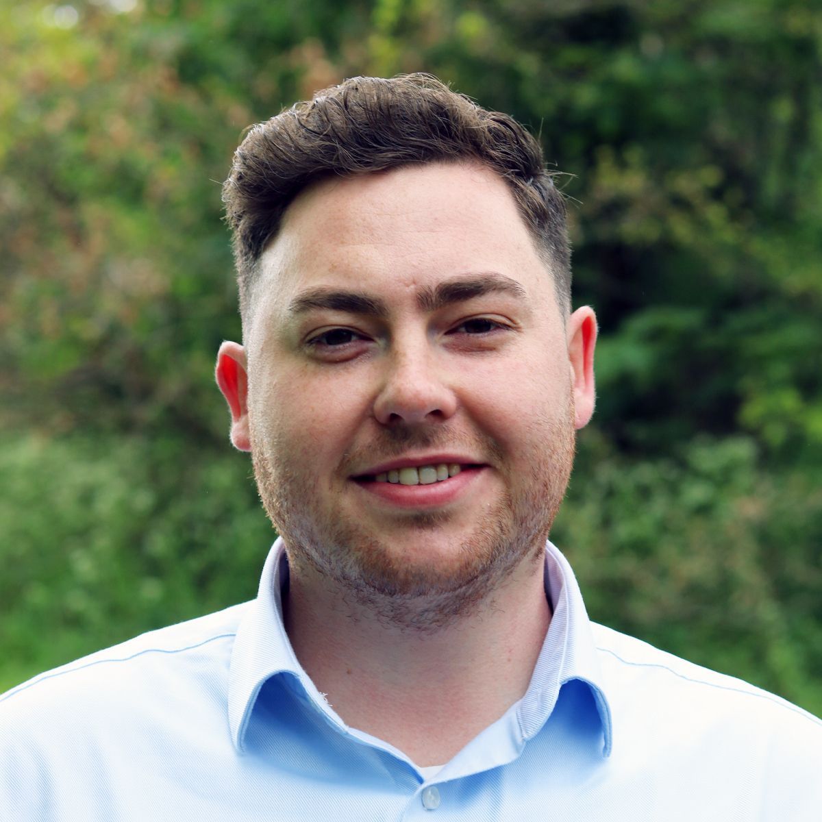 Man in a light blue shirt smiling outdoors with green foliage in the background
