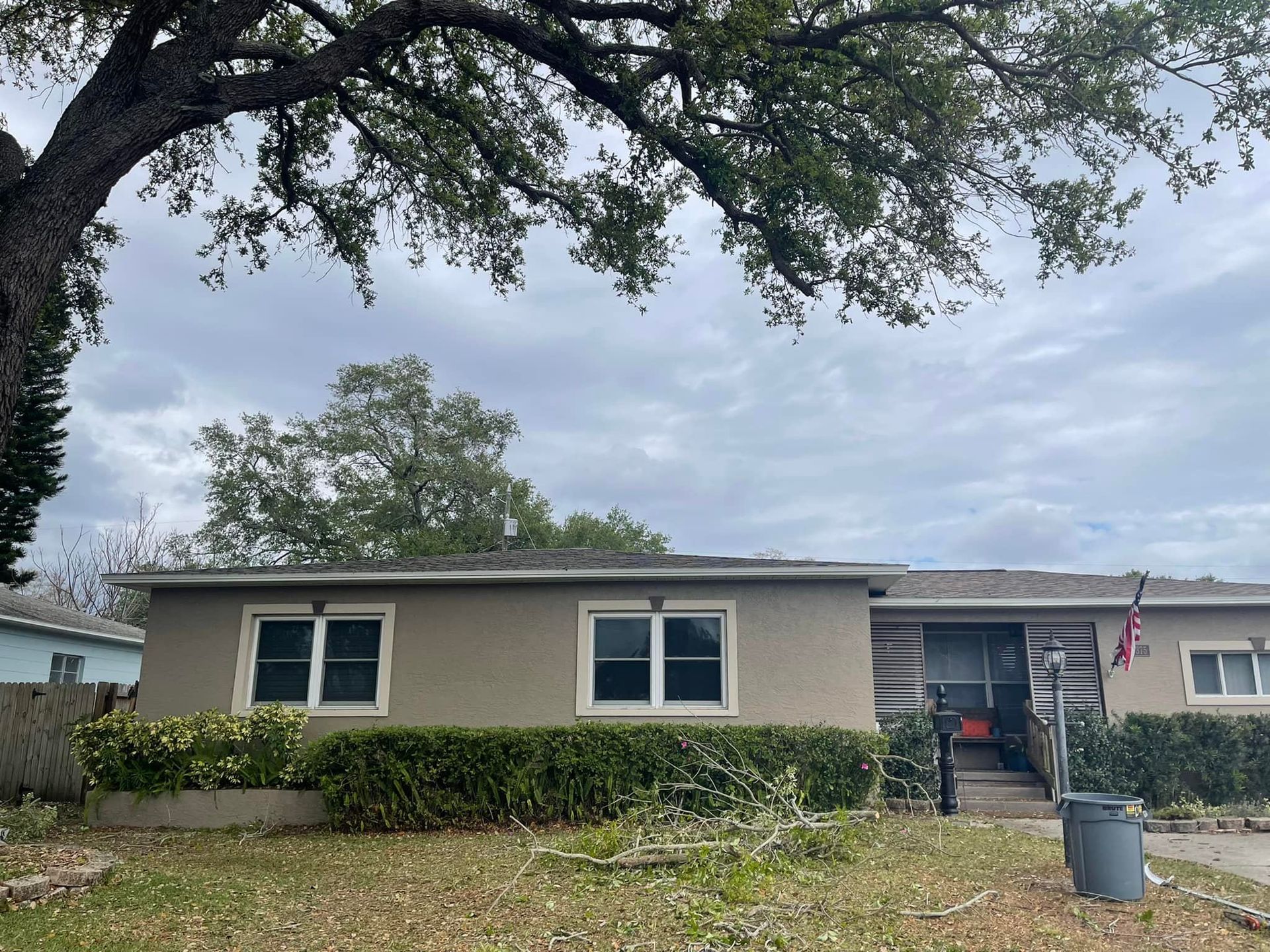 Single-story beige house with a front porch, hedges, and a large tree arching overhead.