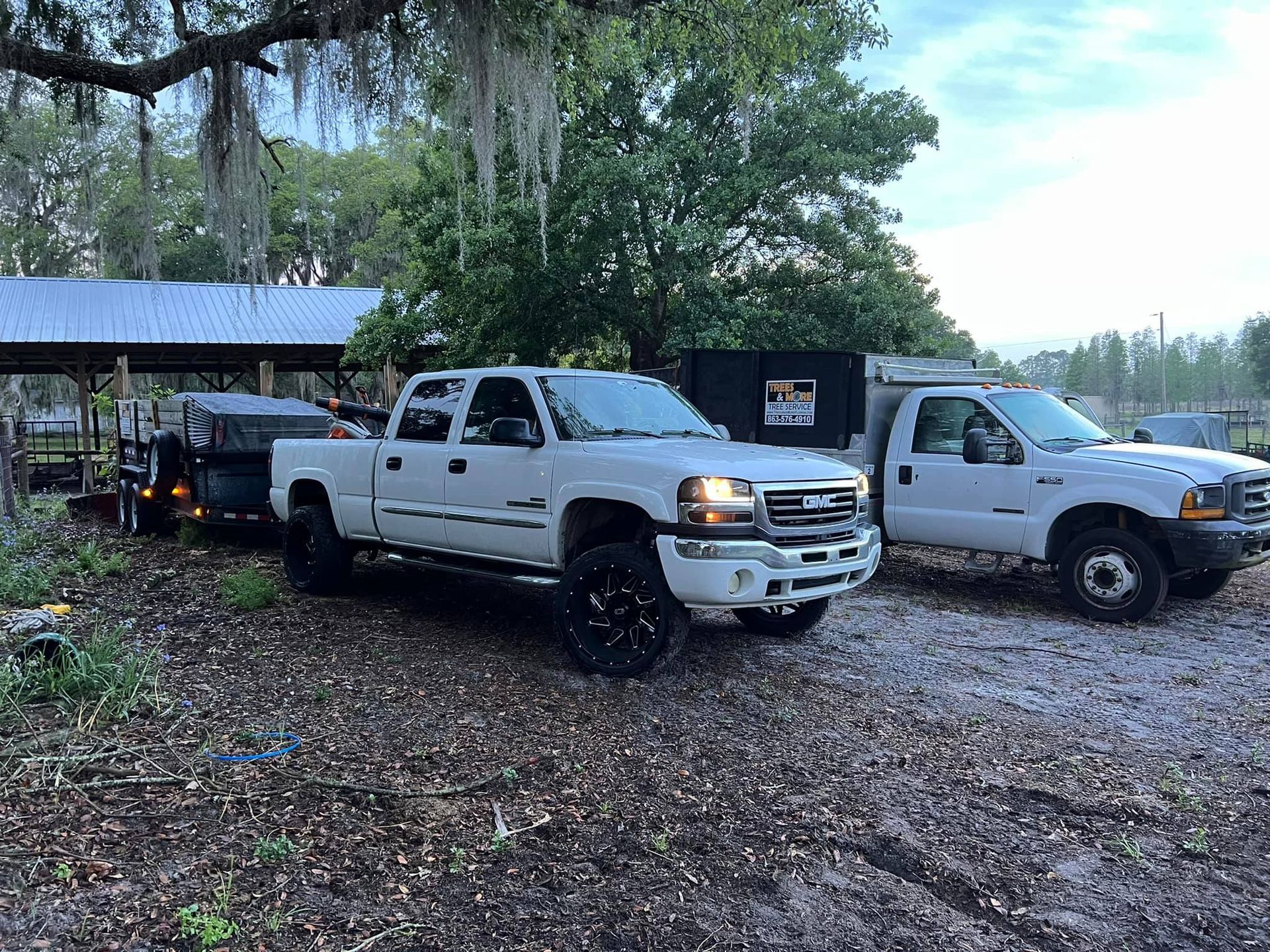 Two white pickup trucks parked on a gravel driveway beside a rural house and trailer in a wooded area