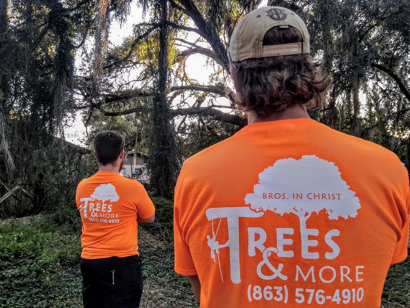 Two people in orange Trees & More shirts standing in a wooded area, viewed from behind.
