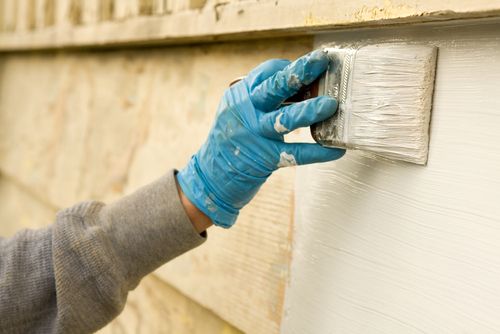 A gloved hand painting the outside of a home an off-white