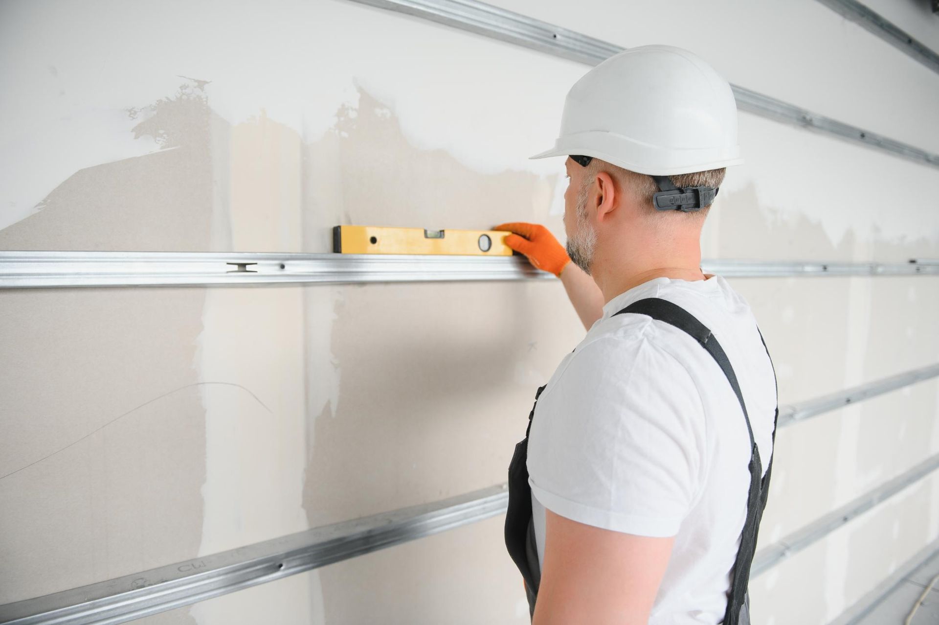 A man in overalls and white hard hat using a level on a half-painted wall