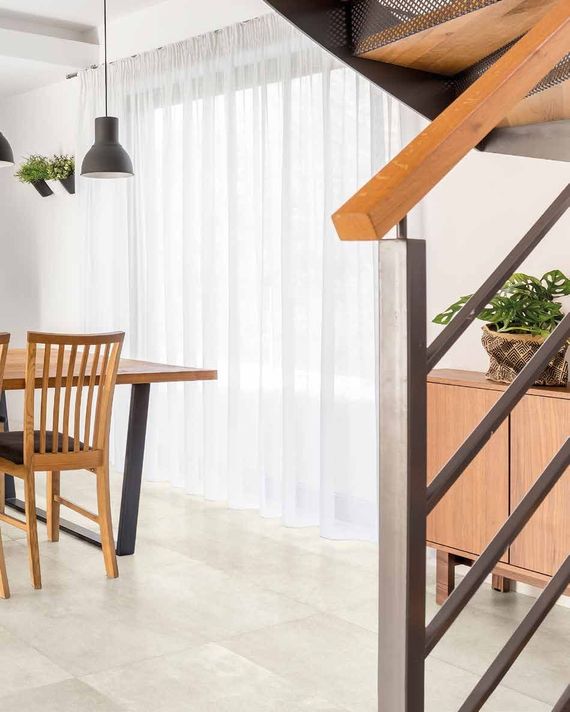 Una zona pranzo con tavolo e sedie in legno, tende bianche trasparenti e una scala con corrimano in legno in primo piano.