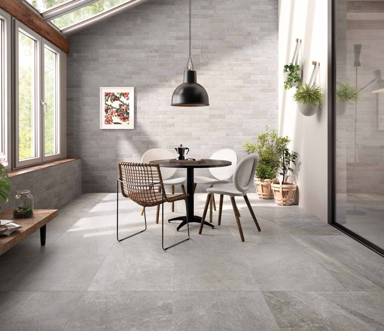 Zona pranzo in veranda con tavolo rotondo, sedie, piante e luce naturale. Pareti e pavimenti grigi.