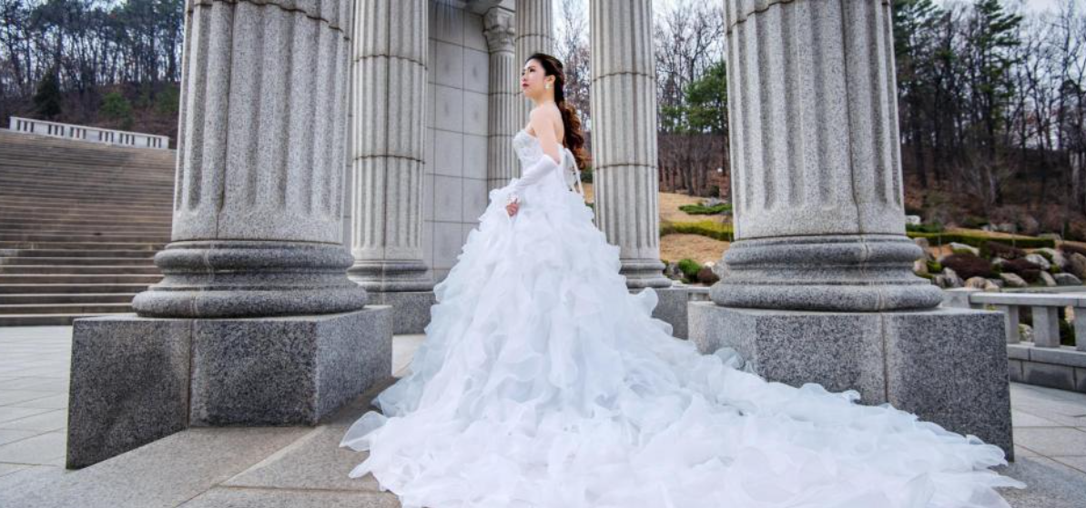 A person in a voluminous white wedding gown stands between large stone pillars in a park-like outdoor setting.
