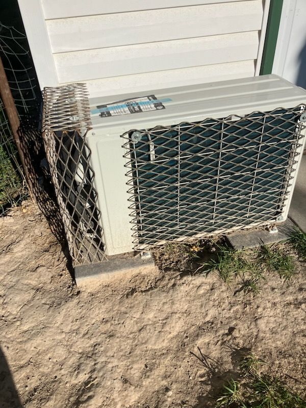 Outdoor air conditioner unit beside a house, enclosed by wire mesh on sandy ground.