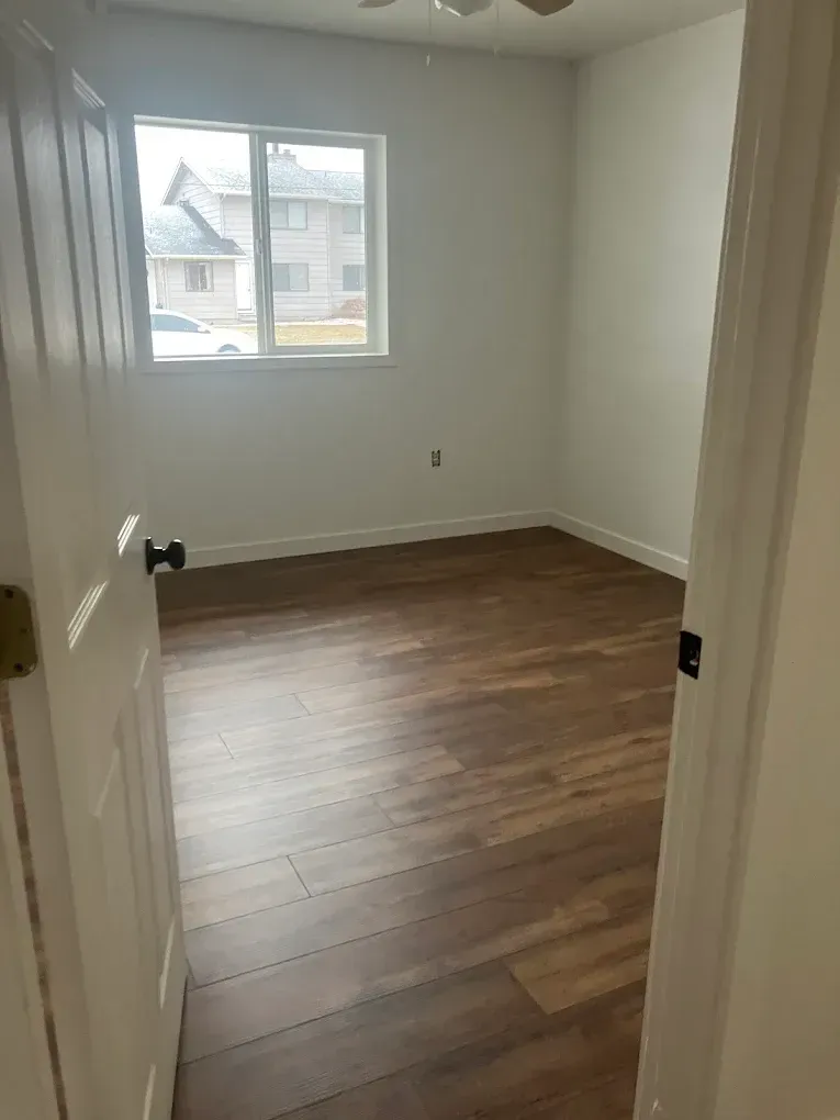 Empty bedroom with wood-look plank flooring, white walls, and a large window, viewed through an open white door.