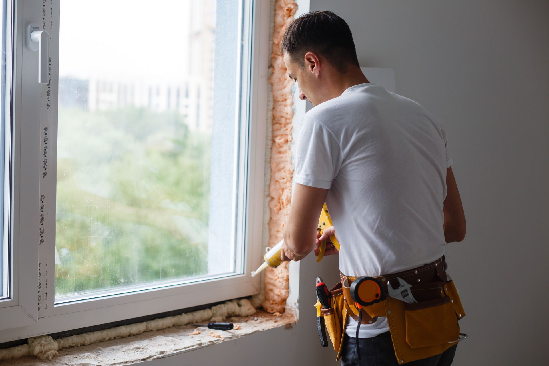 A person in a white t-shirt and tool belt uses a sealant gun to apply foam around the frame of a new window.