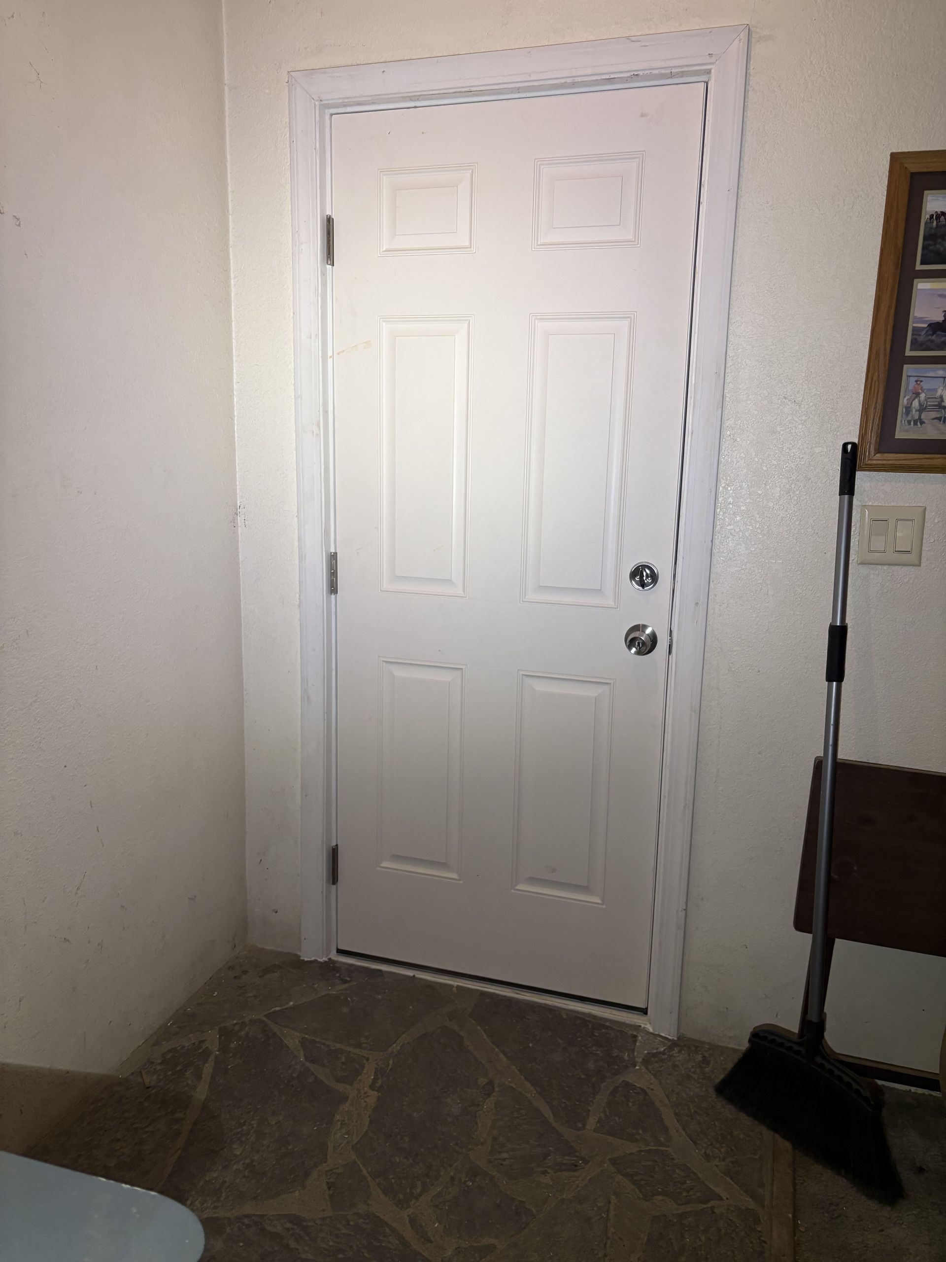 A white six-panel door with silver hardware in a room with a stone floor and a broom leaning against the wall.