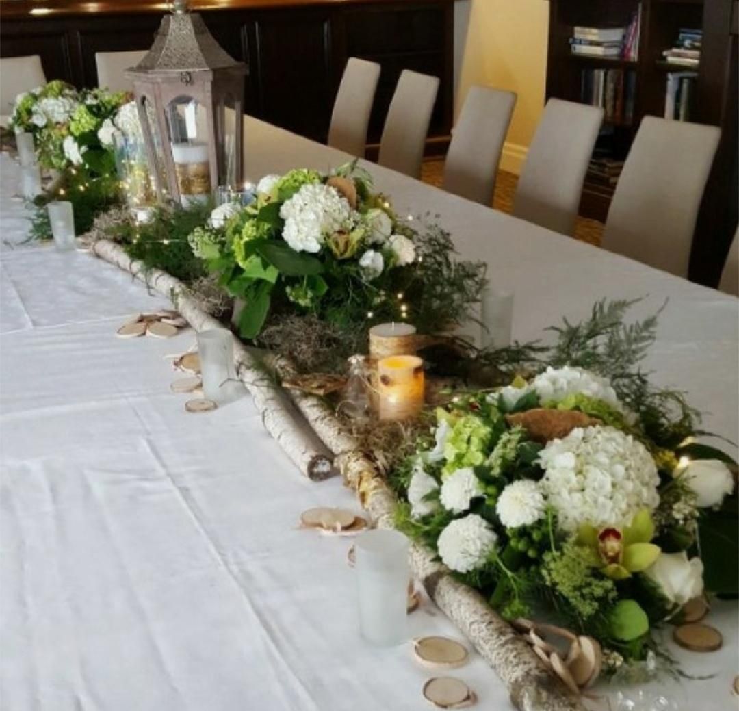 Table centerpiece with white tablecloth, greenery, flowers, candles, and birch logs.