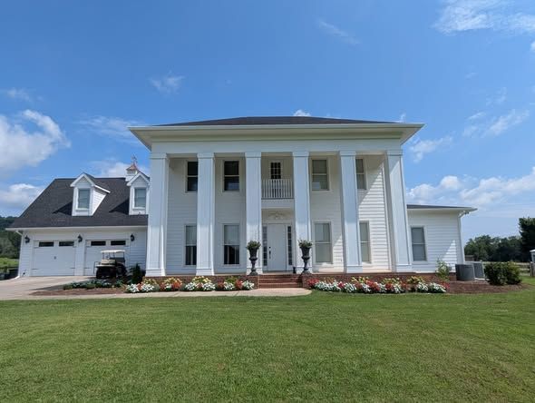 Large white colonial-style house with tall columns, a gray roof, and flower beds in front of a green lawn.