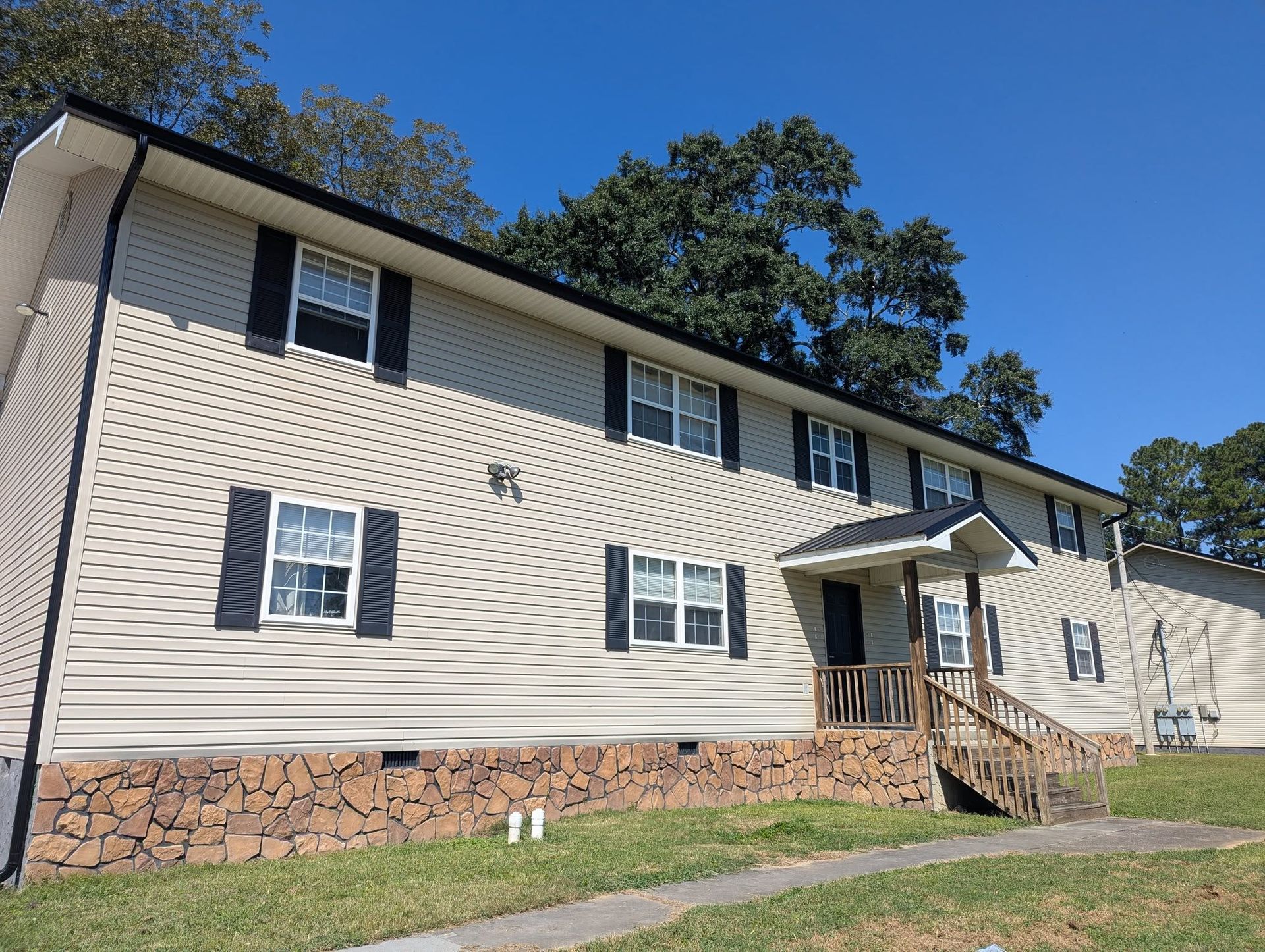 Two-story beige apartment building with dark shutters, stone foundation, and a small front porch under a blue sky