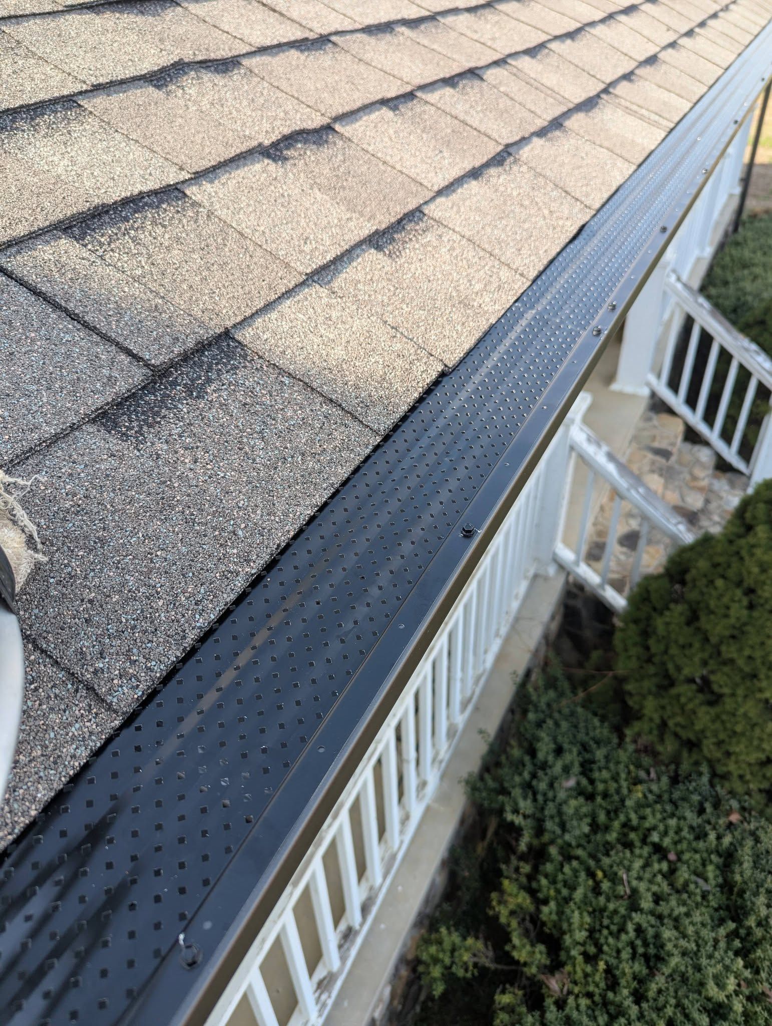 Roof shingles and gutter along a house eave above a white railing and bushes