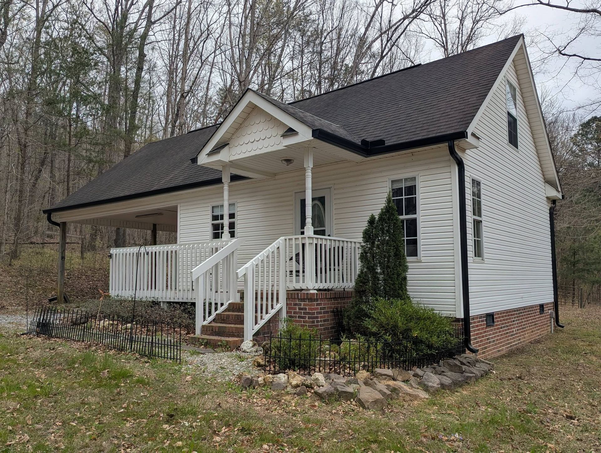 Small white cottage with a front porch, black roof, and brick foundation in a wooded yard