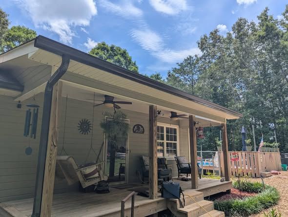 Covered porch with rocking chairs, hanging plants, and potted flowers beside a house under a blue sky