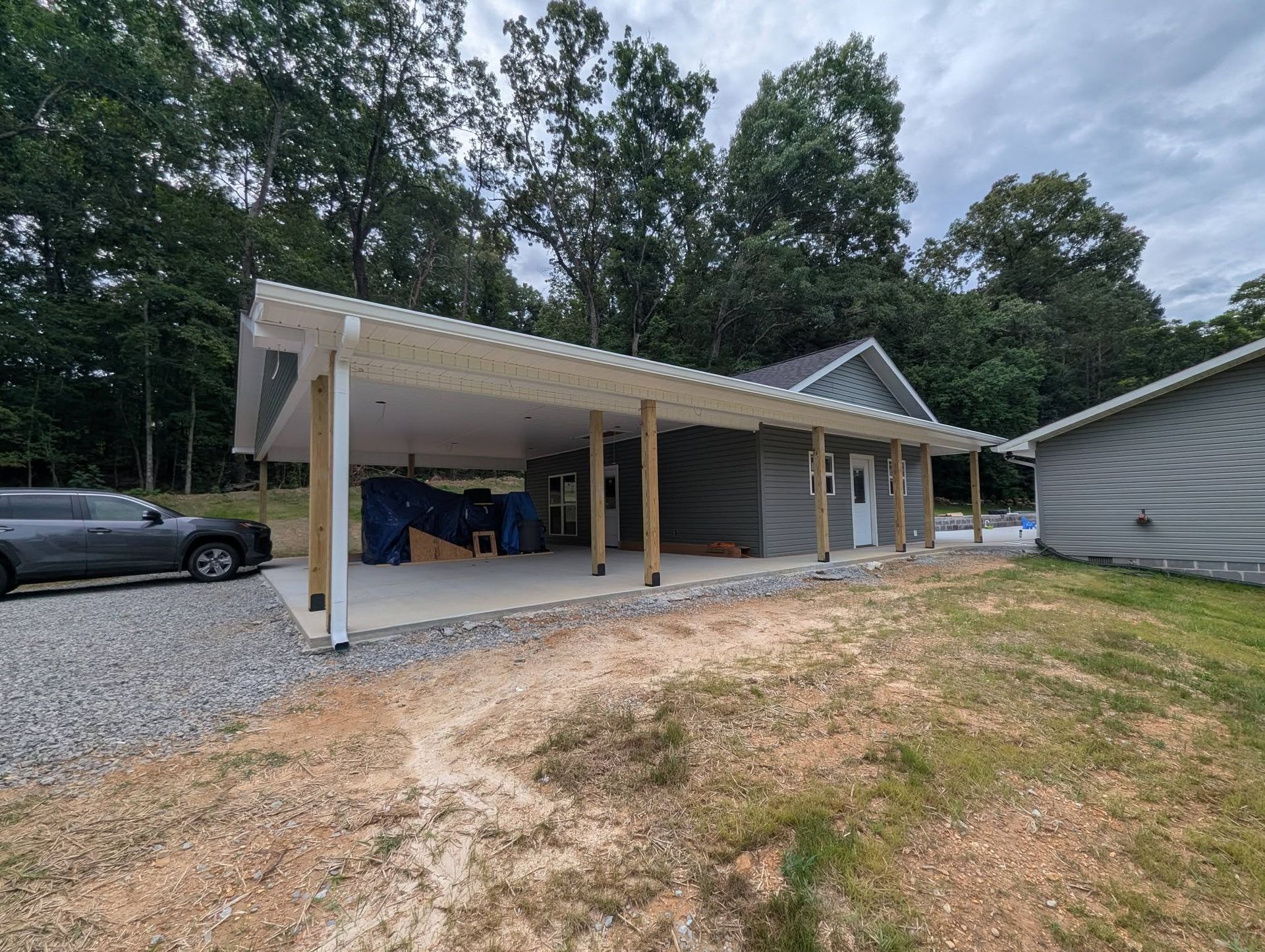 Open-sided carport with a white roof beside a gravel driveway and trees, under cloudy skies