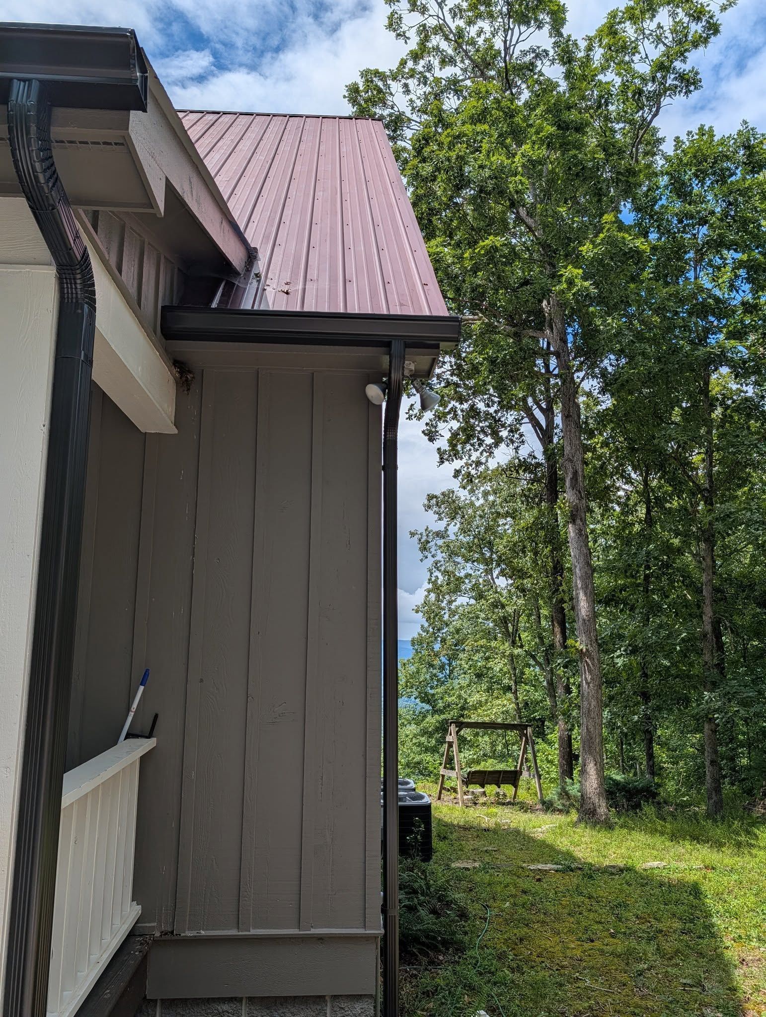 Side of a house with a maroon metal roof beside green trees and grass under a partly cloudy sky