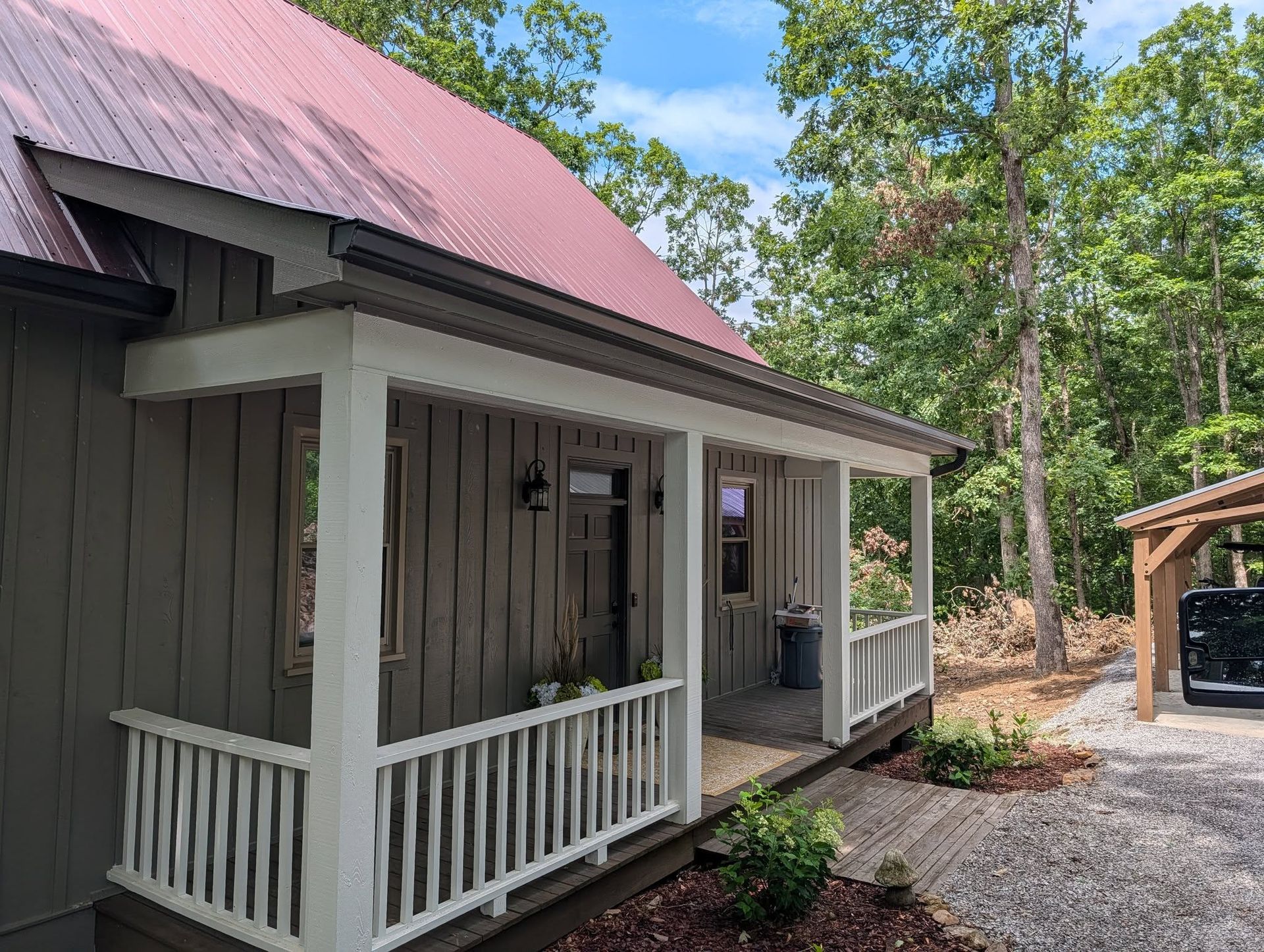 Small gray cabin with a red roof, white porch railings, and a gravel driveway in a wooded area