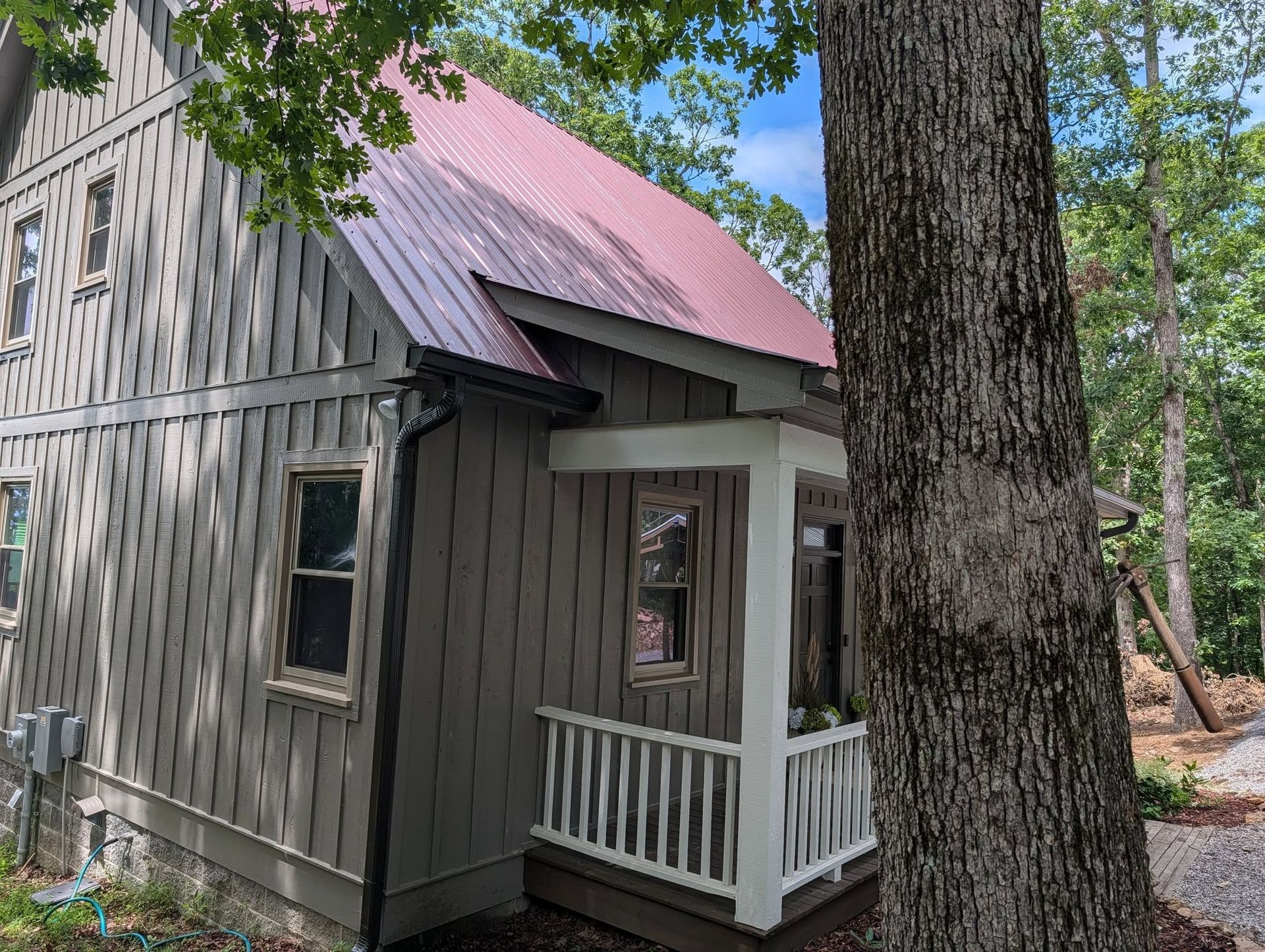 Small gray cabin with a red roof and white porch in a wooded area