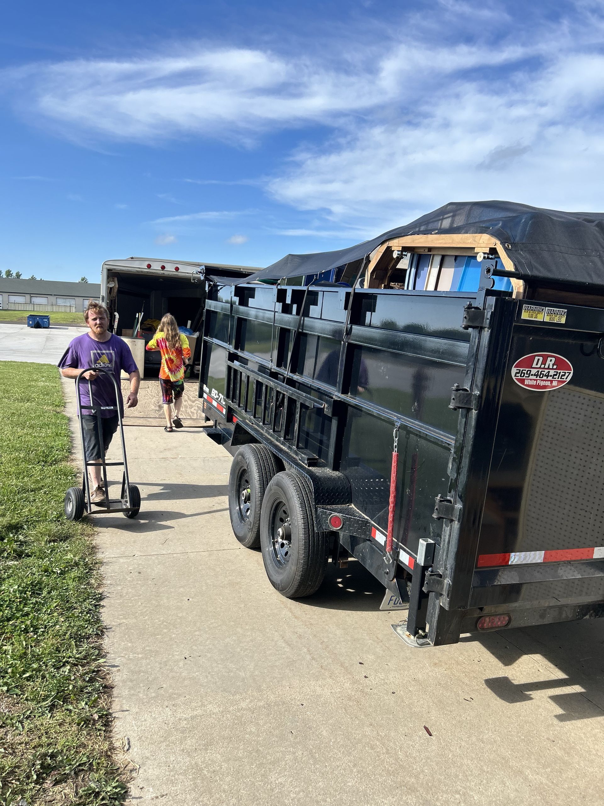 People beside a black dump trailer on a paved path near a lake and mountains.