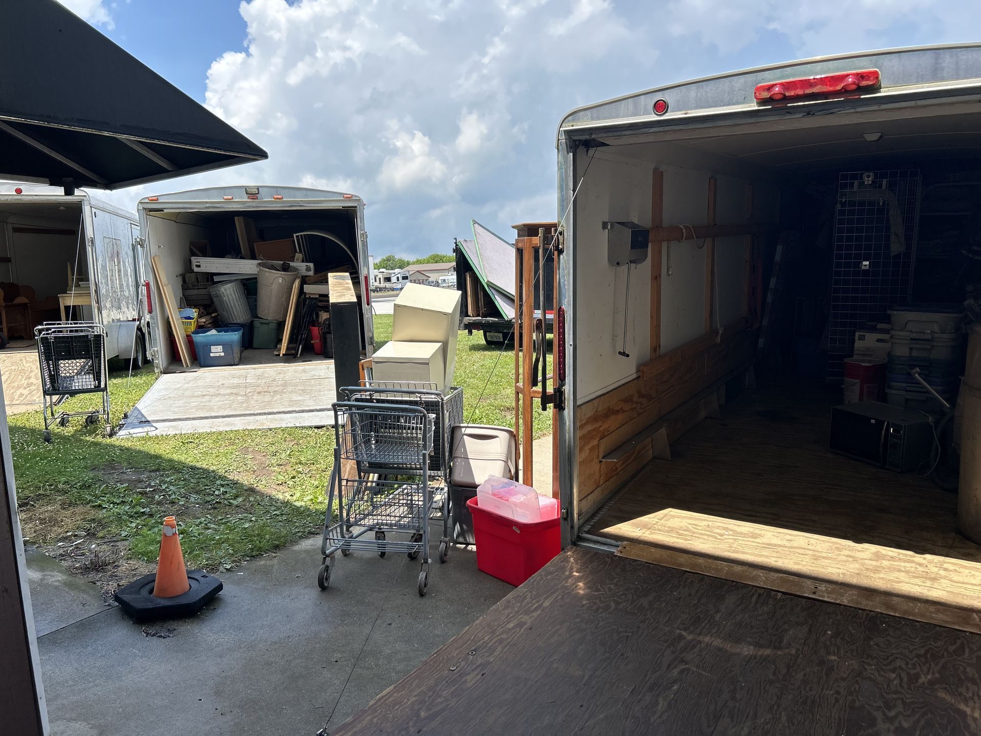 Open truck trailer with a dolly and red cooler in a gravel lot, with tents and another trailer nearby