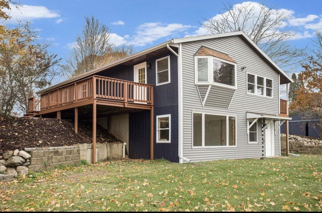 Two-story house with gray siding, dark blue trim, and a wooden deck on a leafy yard under cloudy skies