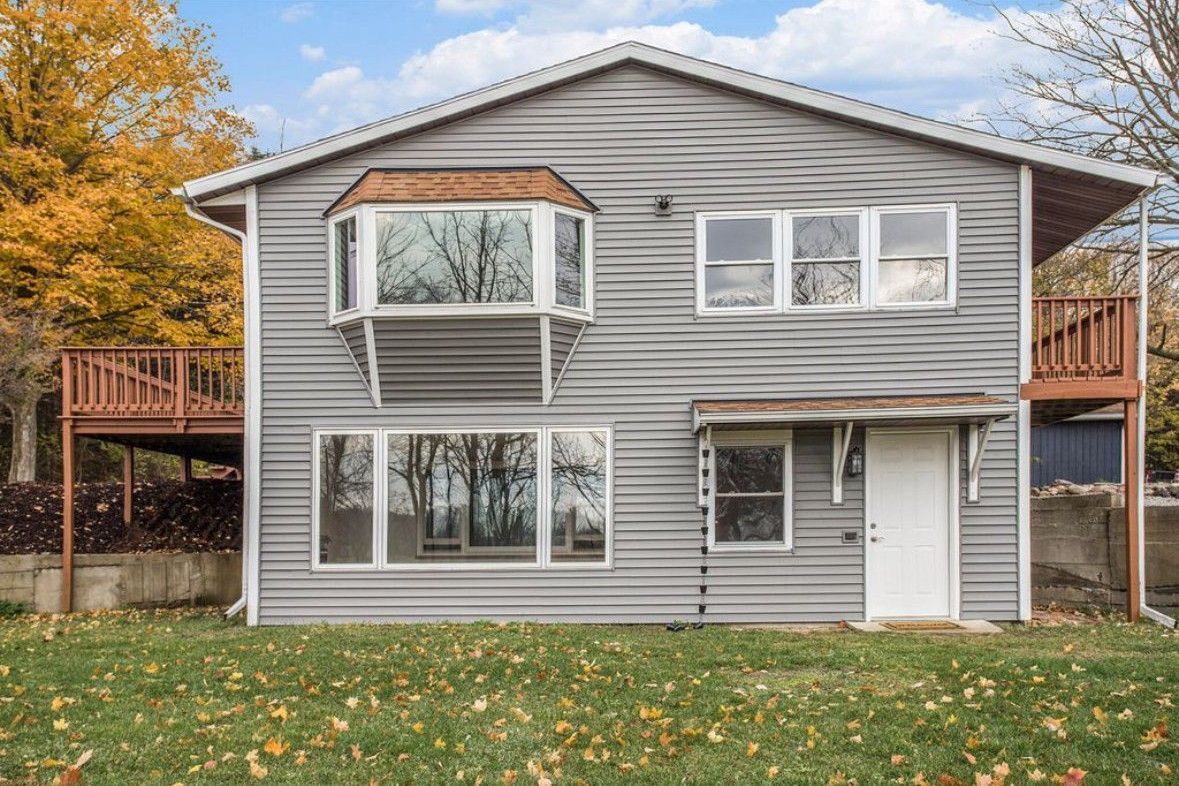 Two-story gray house with white trim and balcony, framed by autumn trees and a leaf-covered lawn