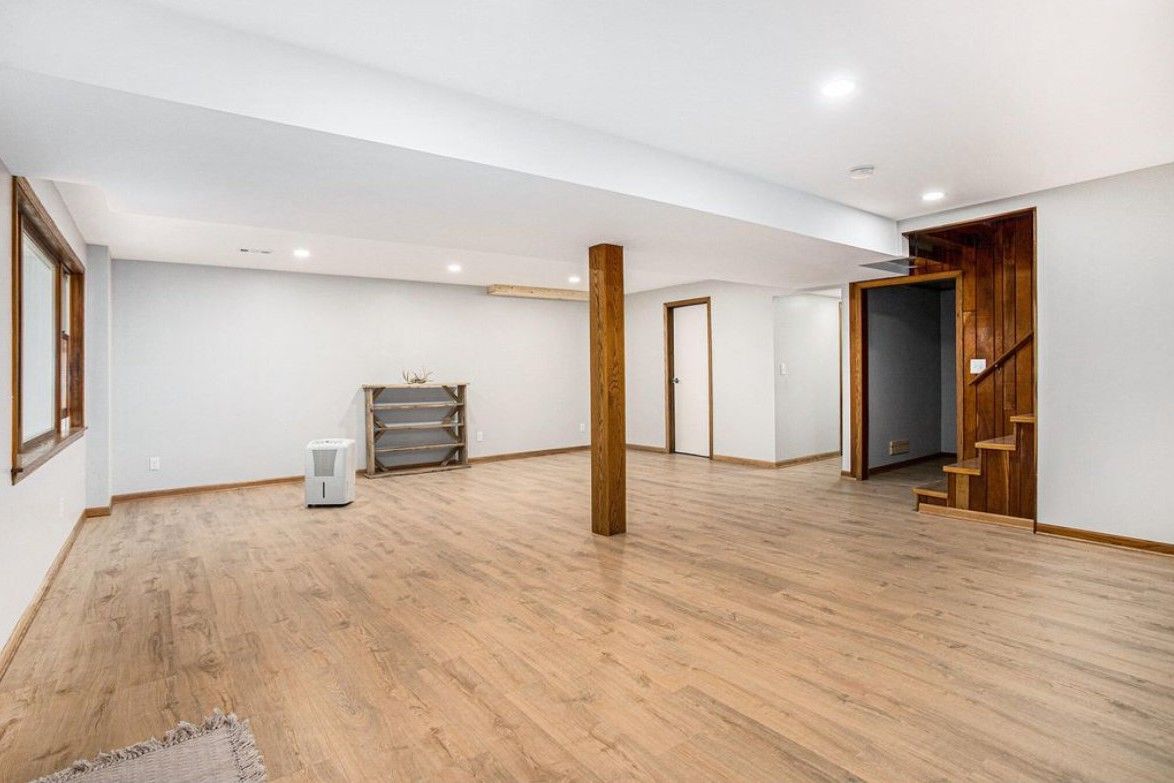 Empty basement room with light wood floors, white walls, exposed wooden post, and recessed ceiling lights