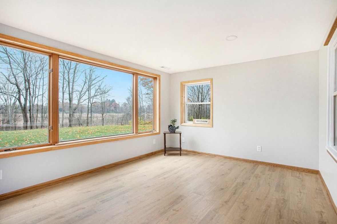Empty sunlit living room with large corner windows, light wood floors, and a small plant table