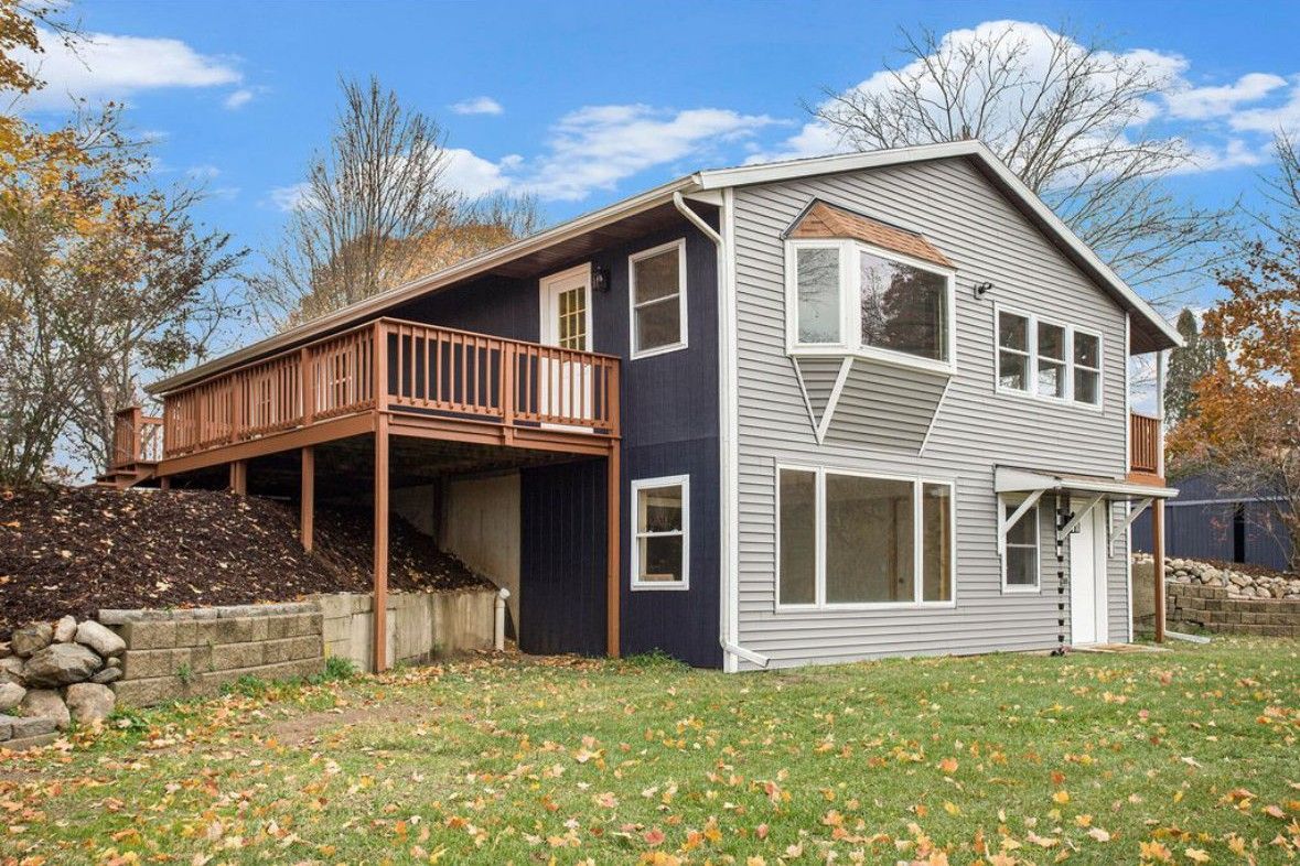 Two-story gray house with a wooden deck and stone retaining wall in a leafy yard under a blue sky