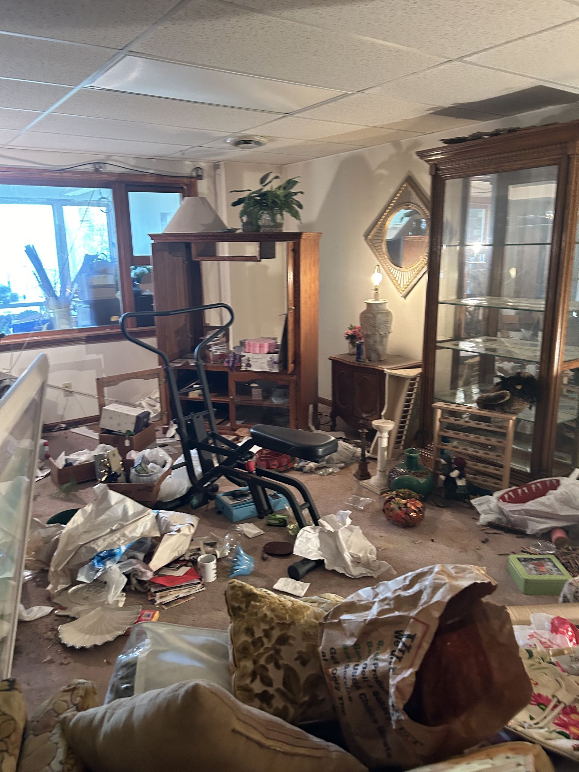 Cluttered living room with overturned furniture, scattered debris, and a broken ceiling panel by a bright window