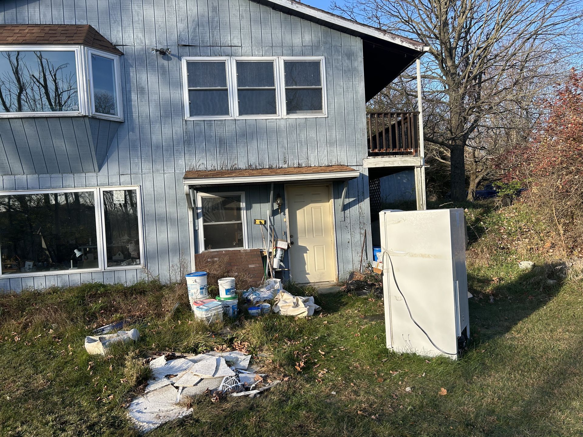 Dilapidated blue house with peeling paint, cluttered yard, and a white appliance standing outside
