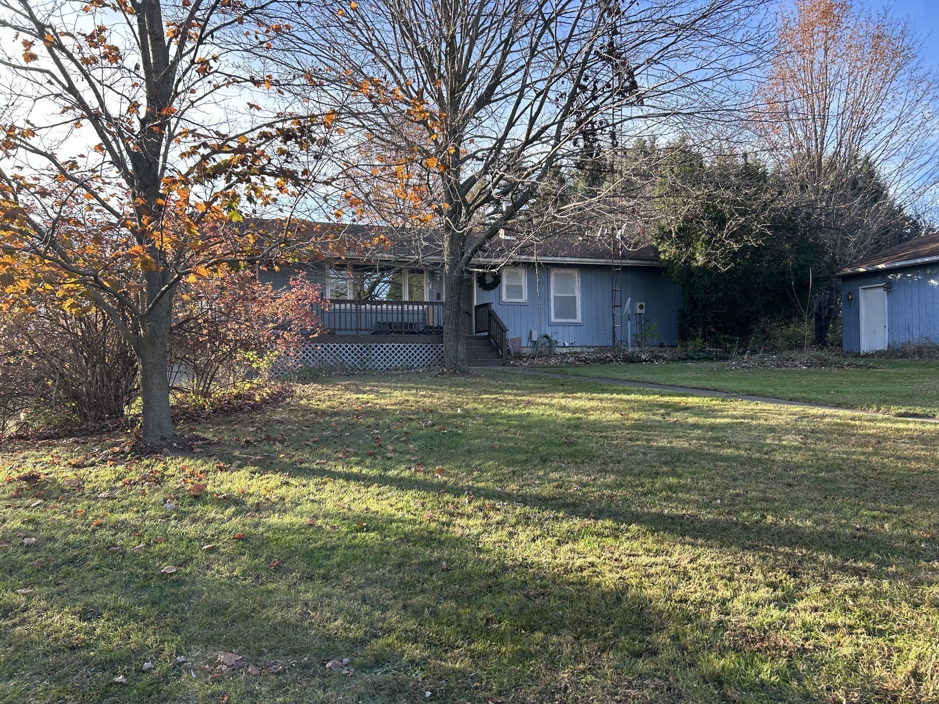 Single-story house with blue-gray siding, large lawn, and trees in autumn sunlight.