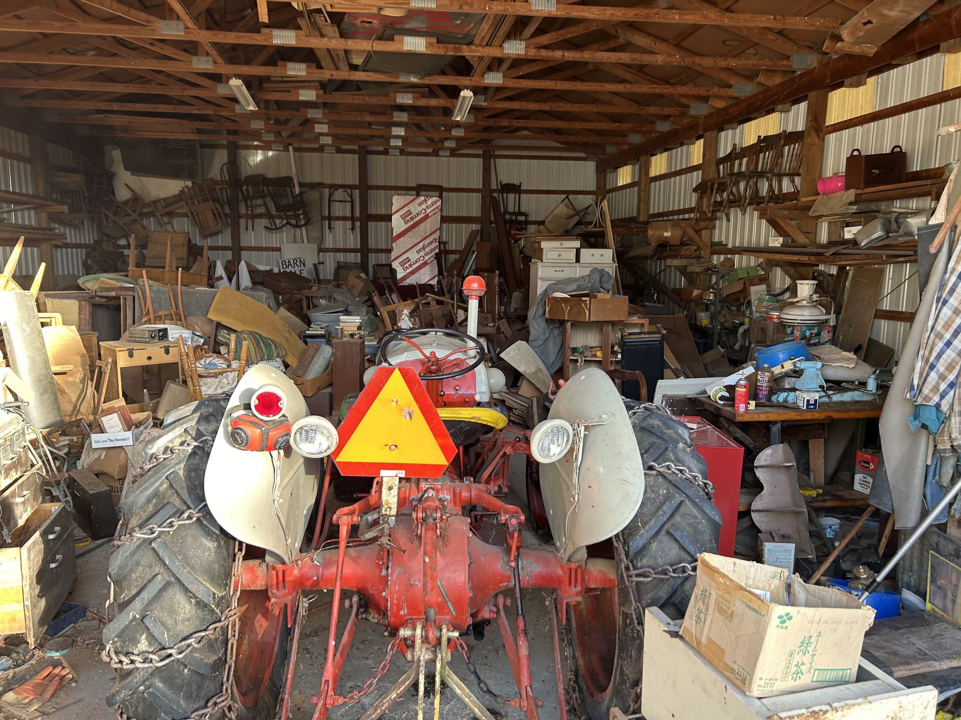Red tractor parked in a cluttered wooden garage with tools, boxes, and equipment stacked around it