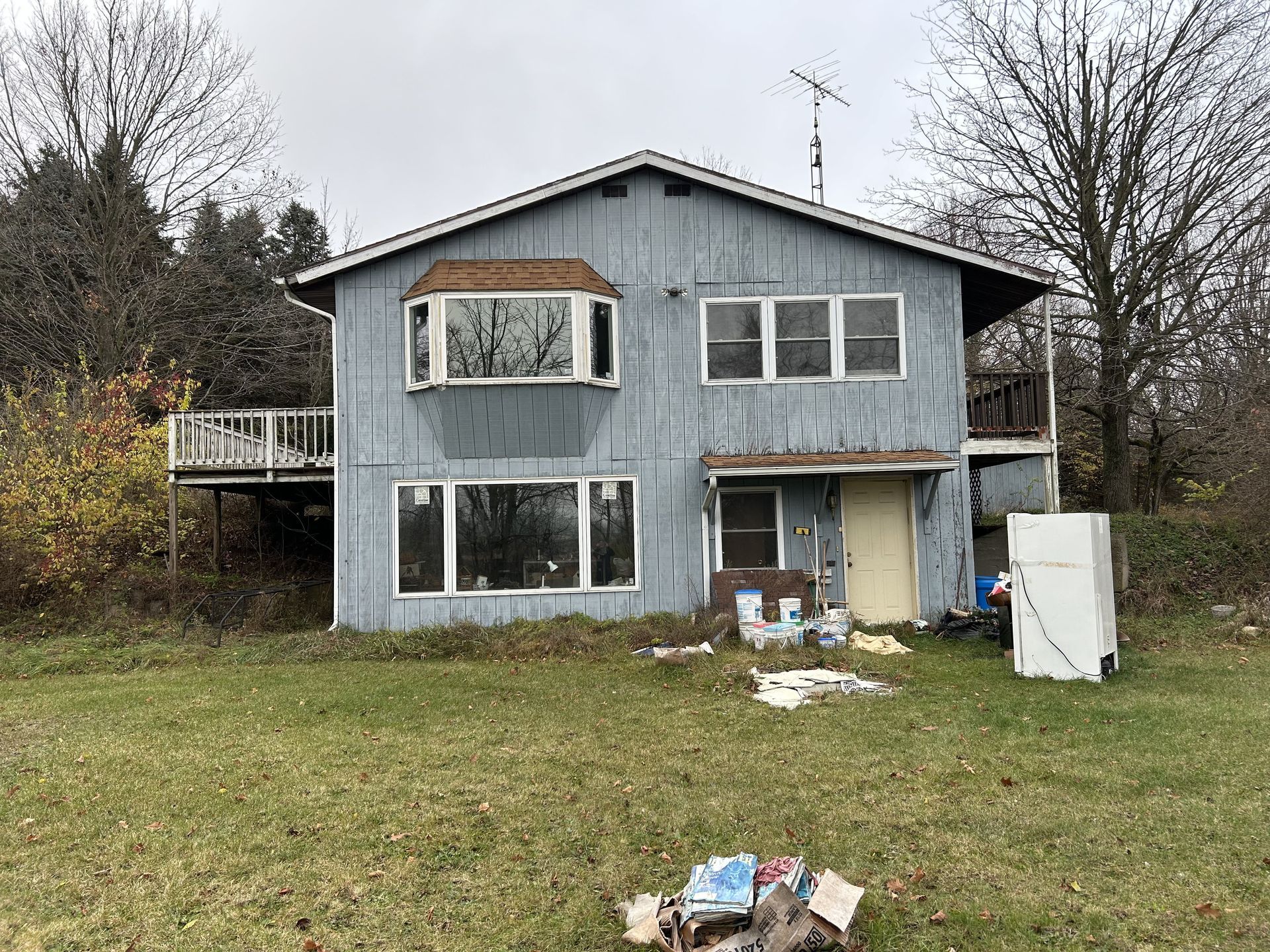 Dilapidated blue-gray two-story house with broken windows, overgrown yard, and scattered trash on the lawn