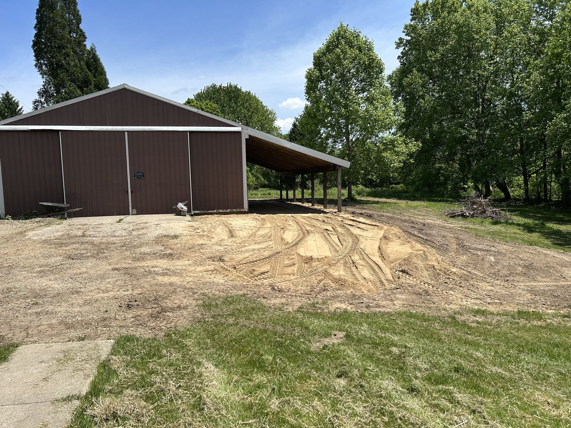 Brown shed beside a dirt lot with fresh tire tracks, surrounded by green trees and grass.