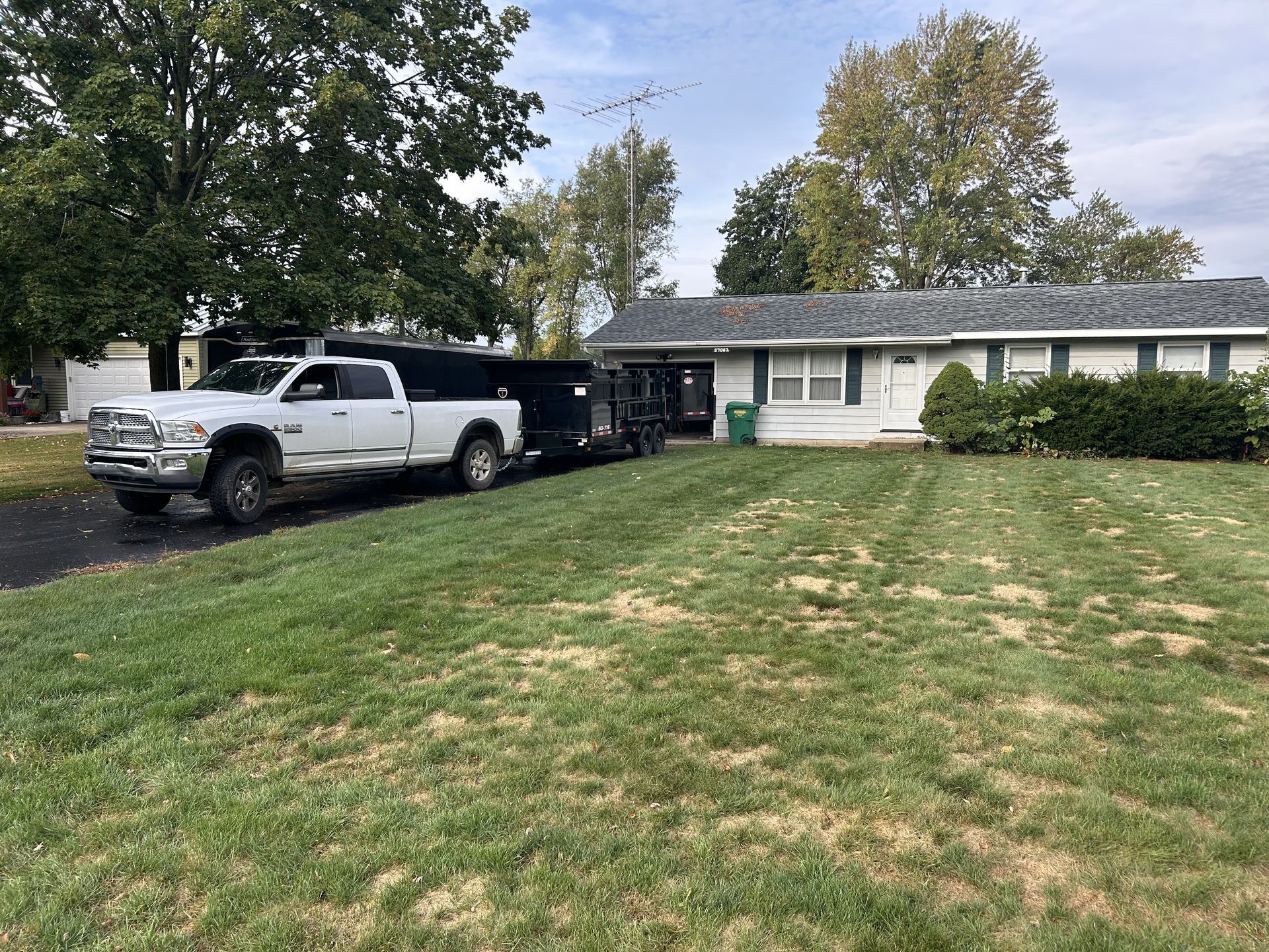 White pickup truck parked on a sloped front lawn beside a single-story house with shrubs and trees.