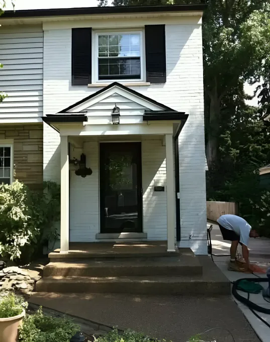 White house with black shutters and a porch, person sweeping the sidewalk.