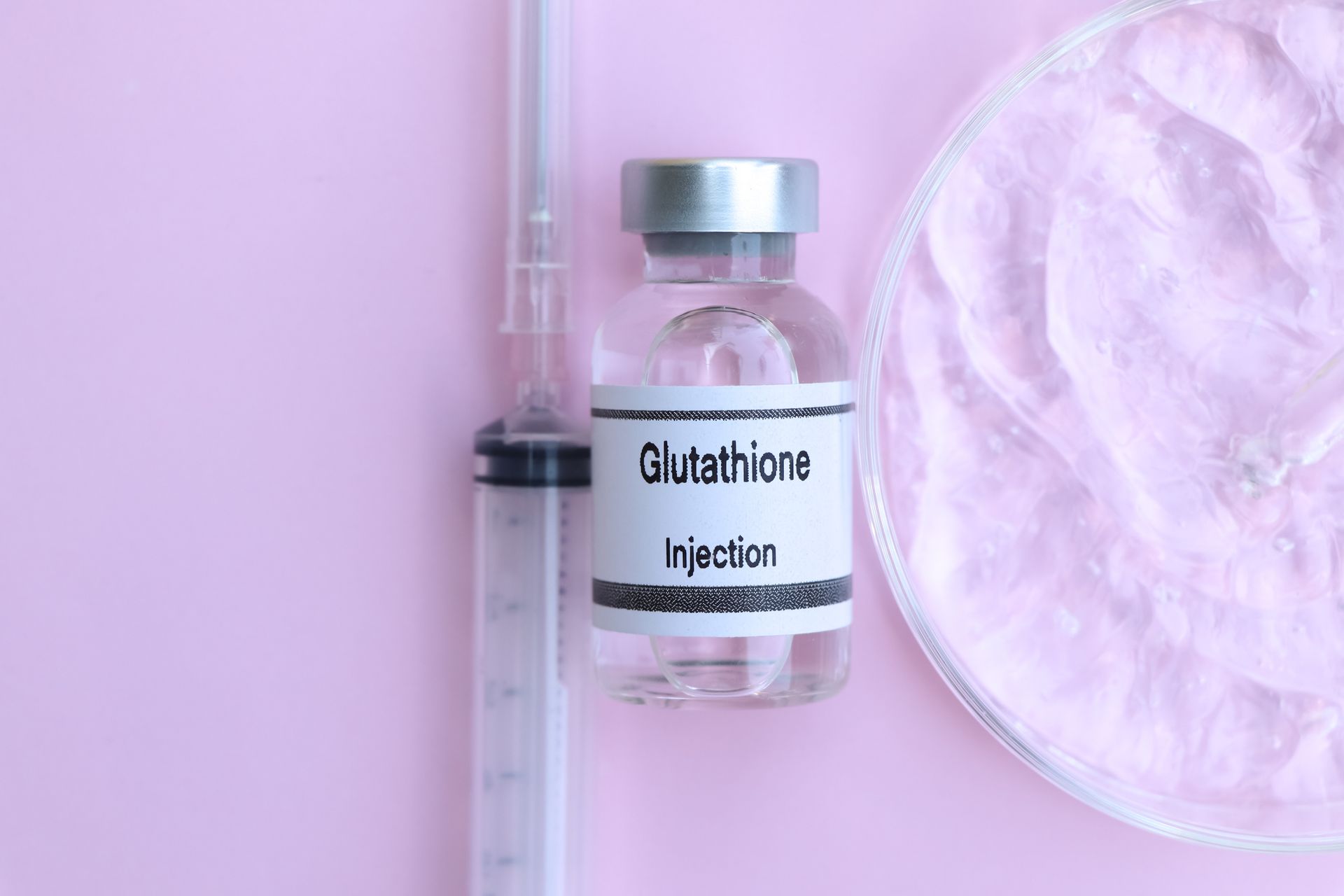 A bottle of glutathione injection next to a syringe on a pink background.