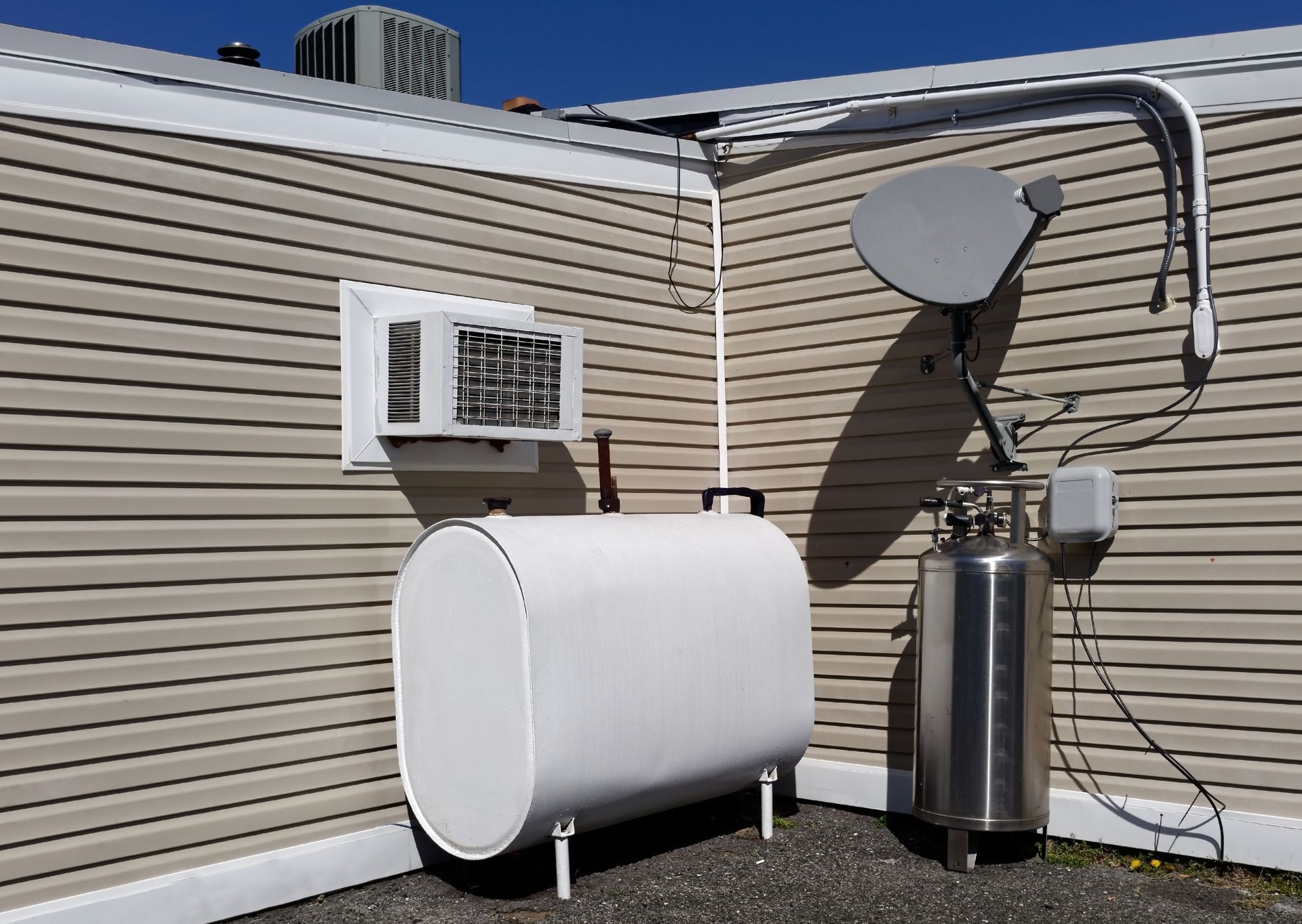 Outdoor HVAC and water-heater setup beside a house, with a window AC unit, white tank, and silver tank.