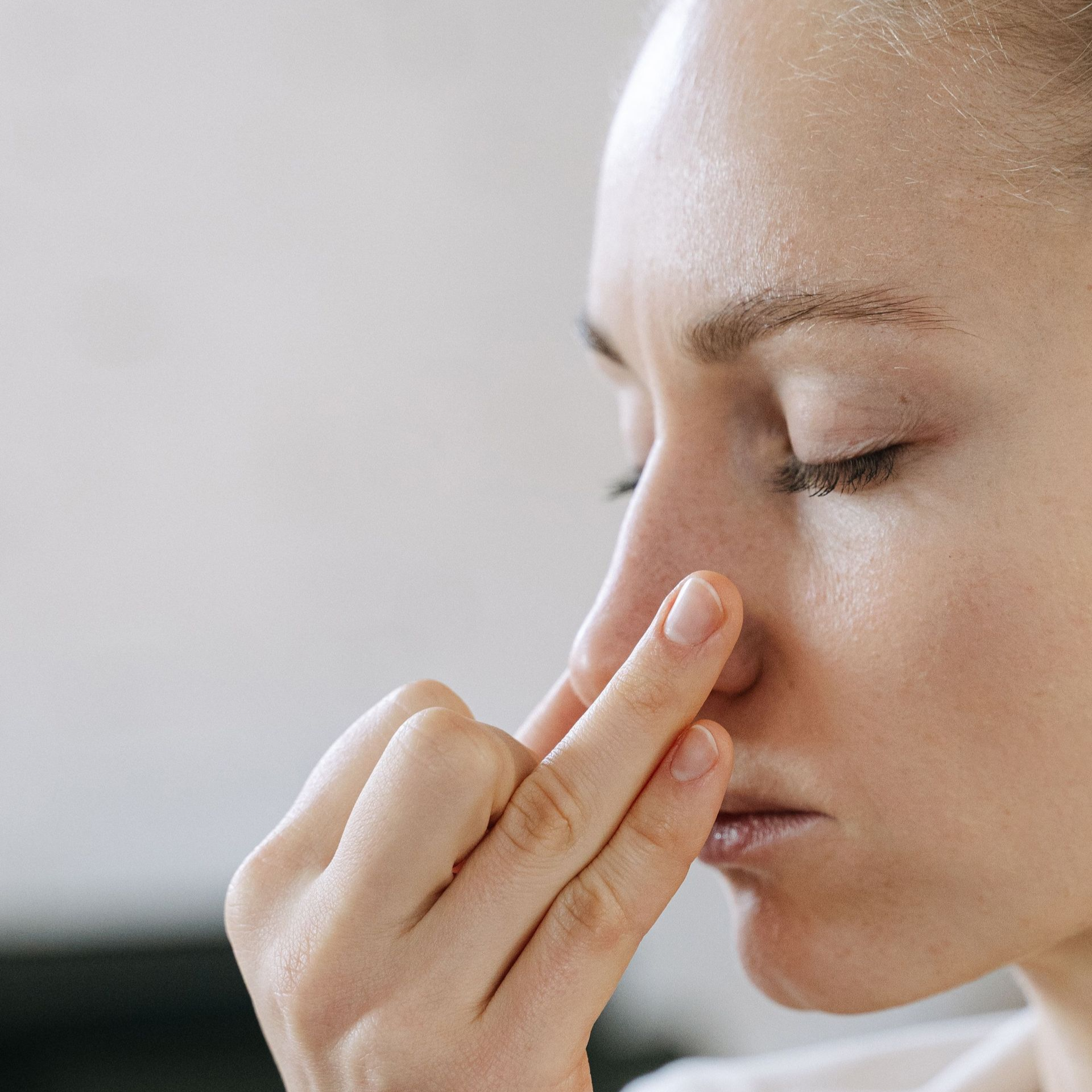 Close-up of a person pinching their nose, eyes closed, against a light background