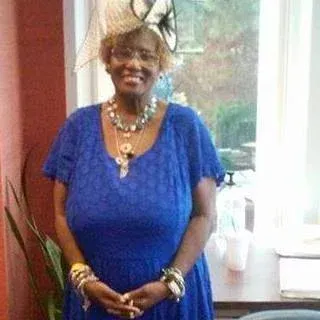 Woman in a blue dress smiling indoors by a window, wearing a large necklace and bracelets.