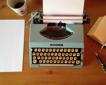 Vintage blue typewriter on a wooden desk with paper loaded, coffee cup, blank paper, and notepad nearby