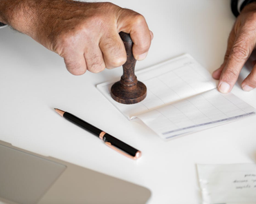 Hands stamping a document on a white desk beside a pencil and papers